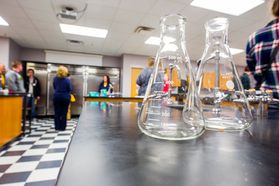 Glass beakers in foreground with students conducting chemistry experiments in background